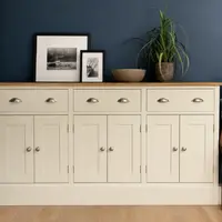 Front view of the cream painted sideboard in a navy blue room, showing the oak top, three drawers with cup handles, six shaker doors with round knobs and a carved wooden armchair alongside