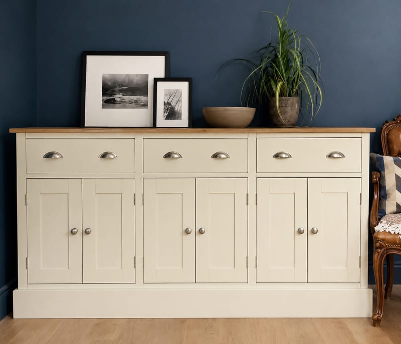 Front view of the cream painted sideboard in a navy blue room, showing the oak top, three drawers with cup handles, six shaker doors with round knobs and a carved wooden armchair alongside