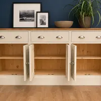 All six cupboard doors opened on the cream painted sideboard, revealing three bays of natural pine with a shelf in each and tongue-and-groove backs, framed prints and a plant on the top