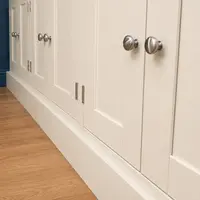 Low raking view along the closed cupboard doors, showing the brushed nickel knobs and butt hinges, the shadow lines of the shaker frames and the plinth meeting the oak flooring
