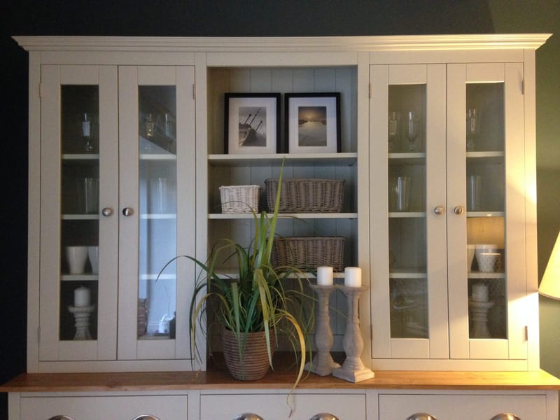 Close view of the dresser top, showing the moulded cornice, glazed doors with glassware inside and the central open shelves holding two framed coastal prints and three wicker baskets
