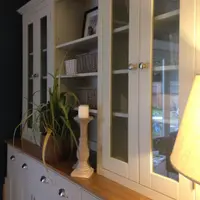 Angled view along the dresser top from the side, showing the glazed doors reflecting a garden, the open shelves with wicker baskets and a candle on a stone pillar beside a trailing plant