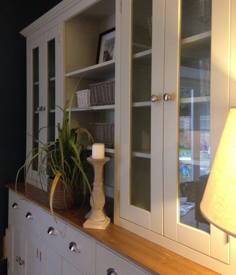 Angled view along the dresser top from the side, showing the glazed doors reflecting a garden, the open shelves with wicker baskets and a candle on a stone pillar beside a trailing plant