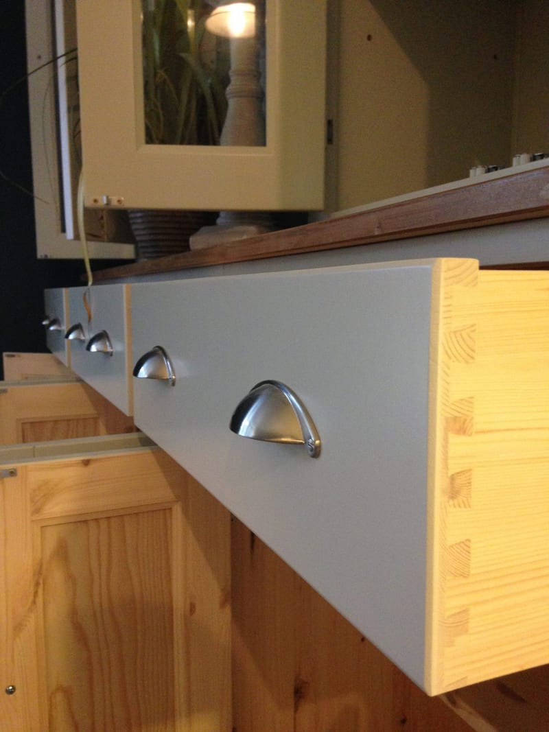 Detail of a drawer drawn fully out of the dresser base, seen side-on so the painted front, brushed nickel cup handle and the pine dovetail joints of the drawer box are all visible