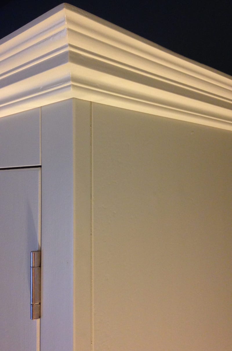 Close detail of the painted cornice at the top corner of the dresser, showing the stepped moulding, the flat side panel and a brushed nickel hinge on the edge of the cupboard door