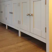 Low view along the base cupboard doors, showing the pairs of brushed nickel knobs, long butt hinges and the square legs the dresser stands on above oak boarded flooring
