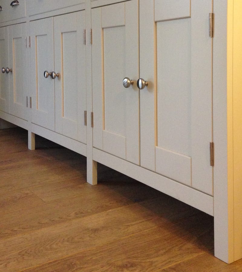 Low view along the base cupboard doors, showing the pairs of brushed nickel knobs, long butt hinges and the square legs the dresser stands on above oak boarded flooring