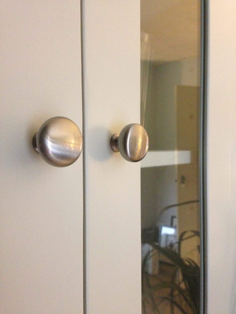 Close-up of two brushed nickel knobs on the meeting stiles of the glazed dresser doors, with the glass panel at the right reflecting the room and a shelf inside the cabinet