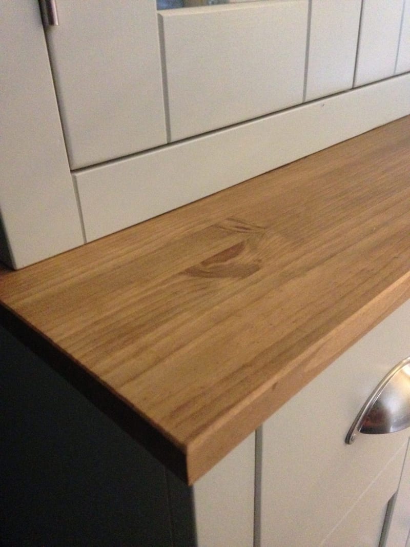 Close view along the waxed pine top of the dresser base, showing the knotted grain and chamfered front edge, the painted rack above and a brushed nickel cup handle to the right