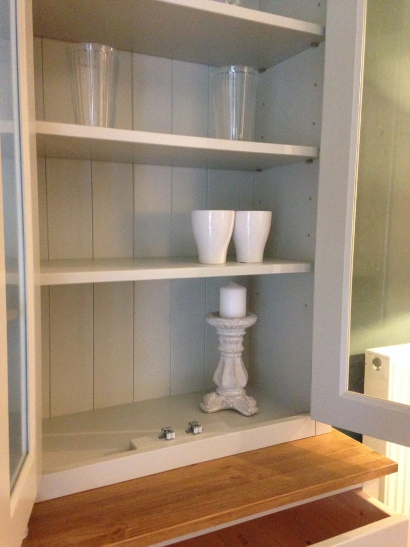 Glazed door opened on the dresser rack, showing the pale blue tongue-and-groove back, three white shelves holding tall glasses, two mugs and a candle on a stone pillar candlestick
