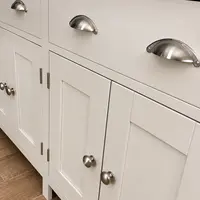 Close view of the drawer fronts and cupboard doors of the cream painted sideboard, showing brushed nickel cup handles, round nickel knobs and the oak top edge above wood-effect tiled flooring