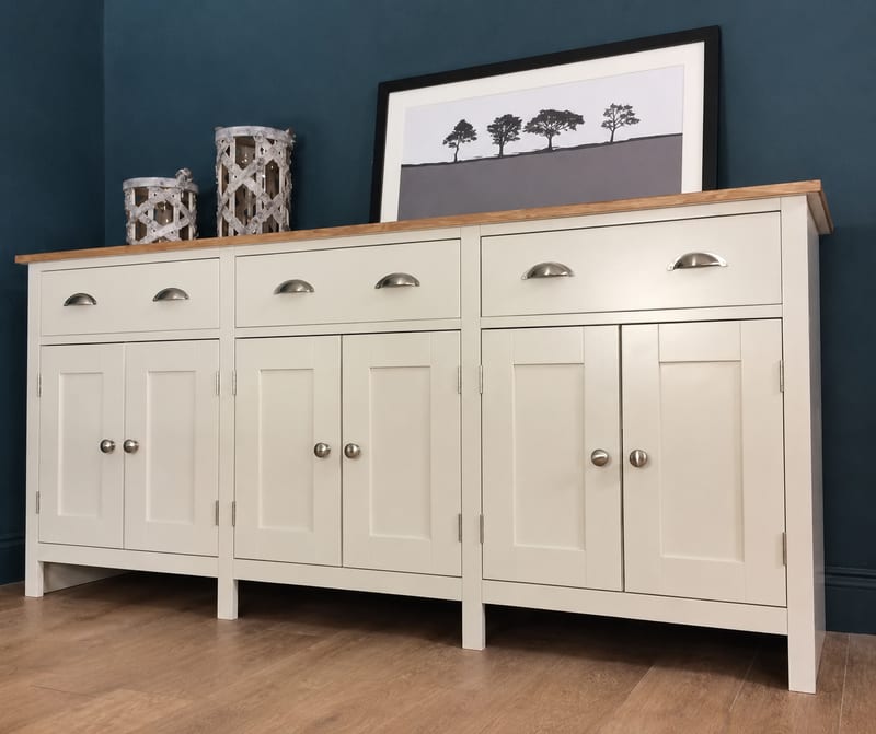 Angled view of a cream painted 6ft sideboard on turned legs, with an oak top, three cup handled drawers and six shaker doors, styled with woven lanterns and a framed tree print against a dark blue wall
