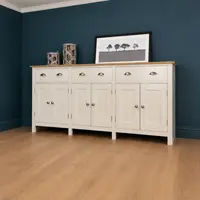 Full view of the cream painted sideboard with oak top against a dark teal wall, six panelled doors and three drawers, topped with two grey woven lanterns and a framed print of trees