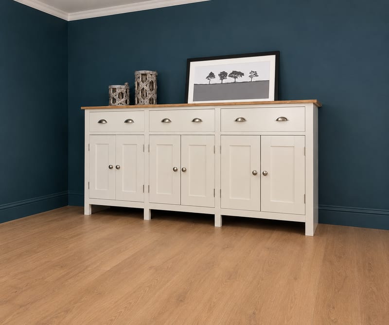 Full view of the cream painted sideboard with oak top against a dark teal wall, six panelled doors and three drawers, topped with two grey woven lanterns and a framed print of trees