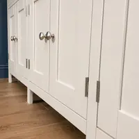 Low angle view along the closed doors of the cream painted sideboard, showing the recessed panels, brushed nickel knobs and butt hinges, the straight square legs and the wood-effect floor beneath
