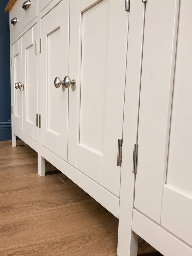 Low angle view along the closed doors of the cream painted sideboard, showing the recessed panels, brushed nickel knobs and butt hinges, the straight square legs and the wood-effect floor beneath
