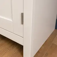 Detail of the end panel and front leg of the cream painted sideboard, showing a brushed nickel butt hinge, the crisp painted edges and the wood-effect plank flooring below