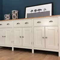 Angled view of a cream painted 6ft sideboard on turned legs, with an oak top, three cup handled drawers and six shaker doors, styled with woven lanterns and a framed tree print against a dark blue wall