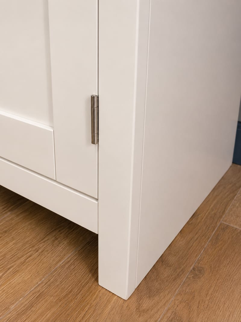 Detail of the end panel and front leg of the cream painted sideboard, showing a brushed nickel butt hinge, the crisp painted edges and the wood-effect plank flooring below