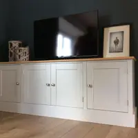 Angled view of the grey painted media unit with oak top and four full height panelled doors with brushed nickel knobs, a television and framed print on top against a dark teal wall