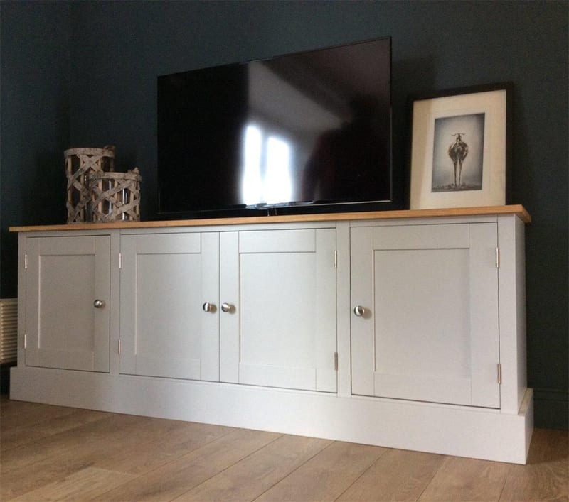 Angled view of the grey painted media unit with oak top and four full height panelled doors with brushed nickel knobs, a television and framed print on top against a dark teal wall