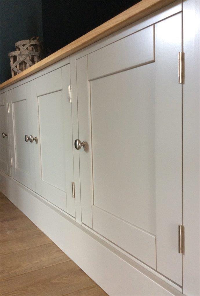 Close angled view along the four painted doors, showing the recessed shaker panels, brushed nickel knobs and long hinges, the oak top above and the deep painted plinth at the base