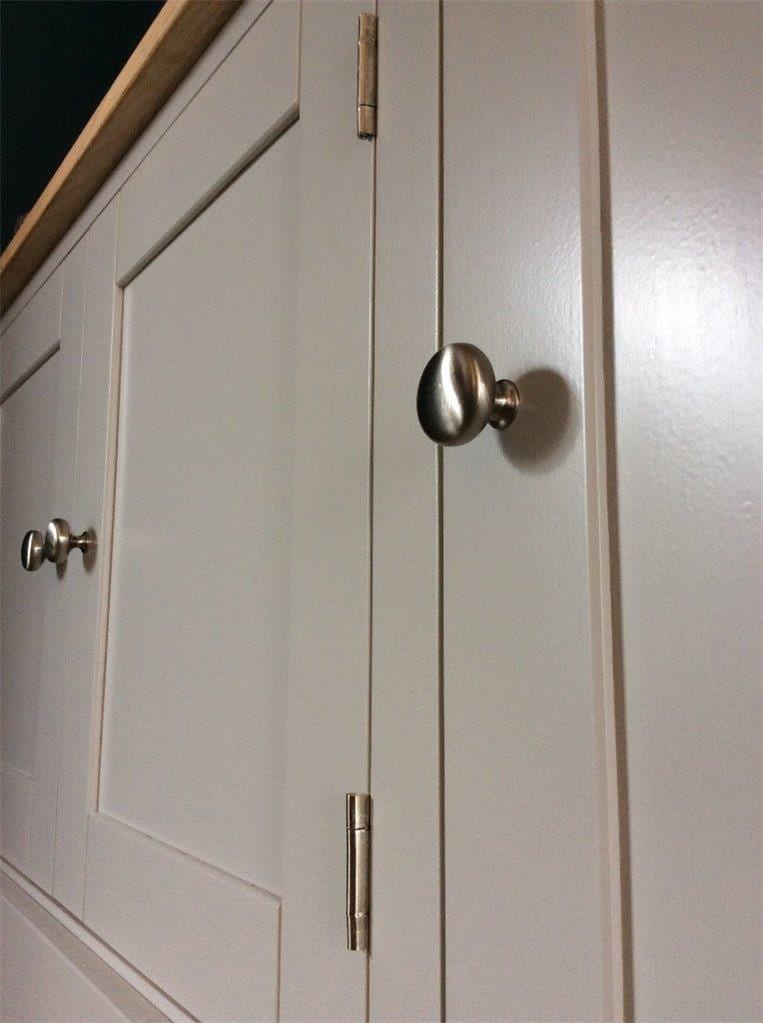 Close view of a round brushed nickel knob on a painted door, with two nickel hinges on the neighbouring door, a further pair of knobs beyond and the corner of the oak top at the upper left