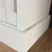 Close view of the bottom corner of the painted media unit, showing the stepped plinth, a brushed nickel hinge on the door edge and the wood plank flooring beneath
