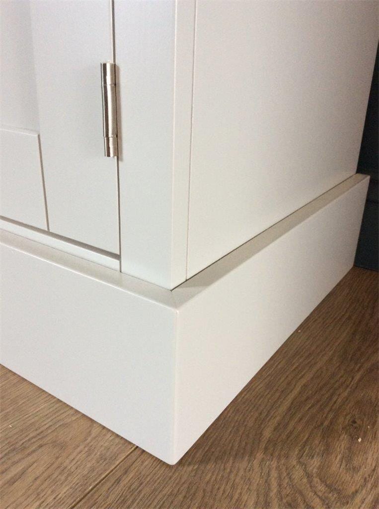 Close view of the bottom corner of the painted media unit, showing the stepped plinth, a brushed nickel hinge on the door edge and the wood plank flooring beneath