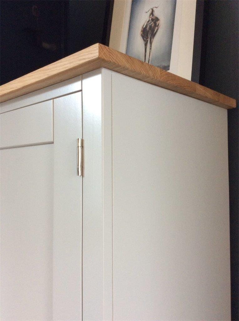 Detail of the top corner of the painted media unit, showing the square planed edge and grain of the solid oak top, a nickel hinge on the door below and a framed print resting on the top