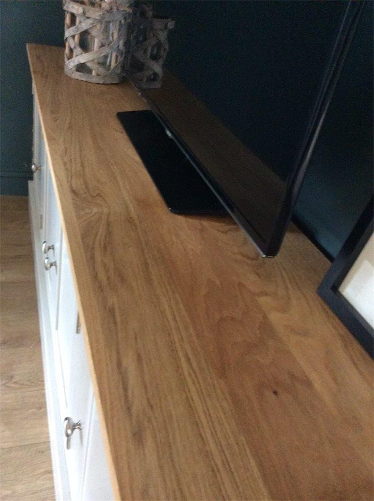 Close view along the solid oak worktop, showing the grain and figure of the boards, the base and stand of the television, a woven lantern behind and four nickel knobs on the doors below