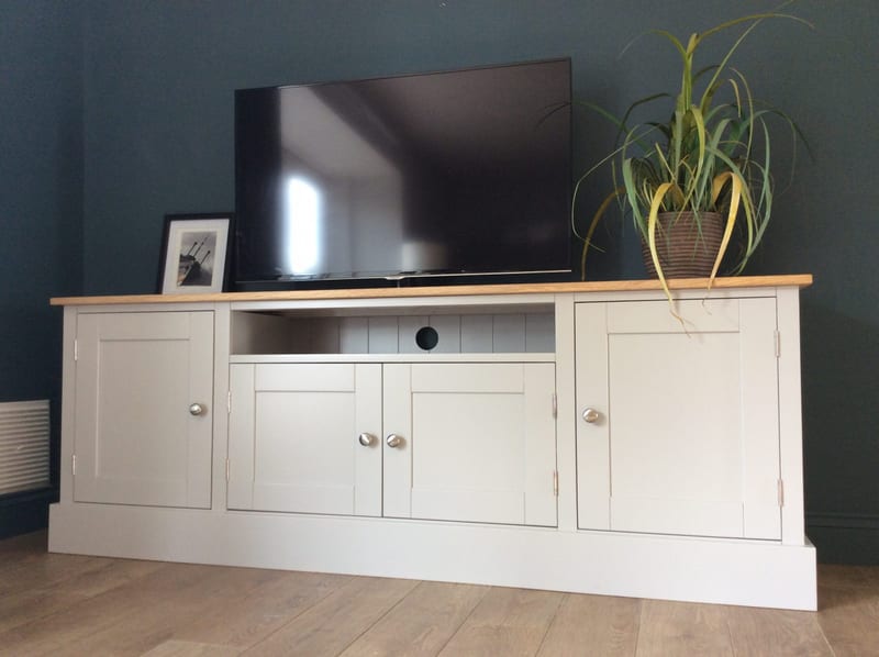 Cream painted 6ft TV media base unit with a solid oak top, an open media shelf with a cable hole between two cupboards, and a television and a plant on top