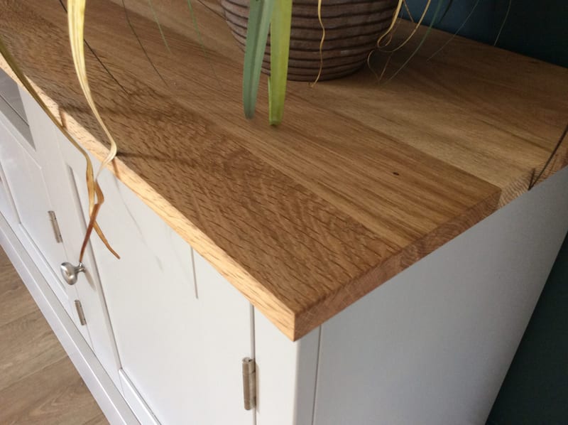 Close view of the oak worktop corner of the painted media unit, showing the sawn edge, prominent grain and a ribbed plant pot, with a single nickel knob and two hinges on the door below