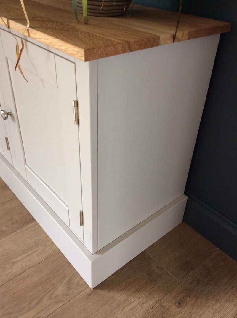 Angled view of the painted end panel and stepped plinth of the media unit, with the oak top overhanging above, two brushed nickel hinges on the door and plank flooring below