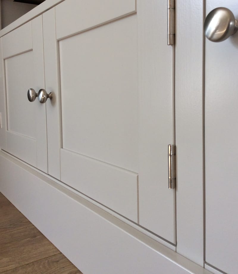 Detail of two painted doors meeting at the centre of the media unit, showing a pair of round brushed nickel knobs, two nickel hinges on the door edge and the recessed shaker panels above the plinth