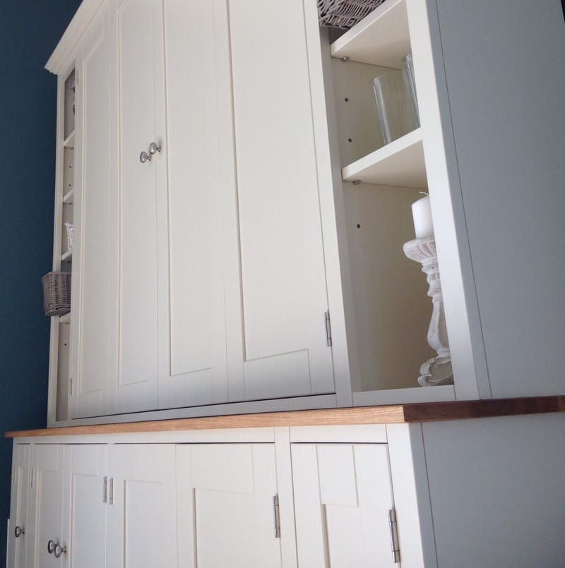 Angled view of the upper section with the doors closed, showing the open end shelves holding a glass vase, a wicker basket and a white candle on a stone candlestick above the oak worktop