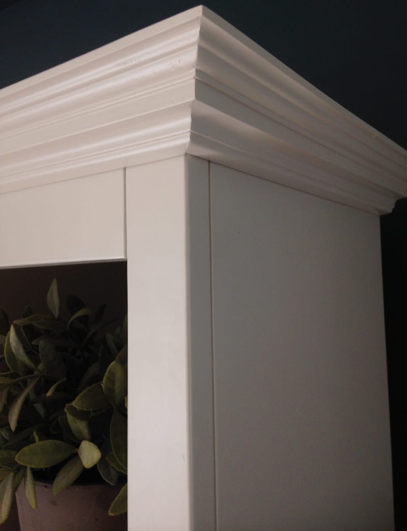 Detail of the top corner of the painted dresser against the dark blue wall, showing the layered moulded cornice, the mitred return and the leaves of a potted plant on the shelf below