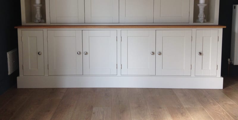 Front view of the base cupboards, six painted shaker doors with round brushed nickel knobs under the oiled oak worktop, a deep plinth on oak boards and stone candlesticks on the shelves above