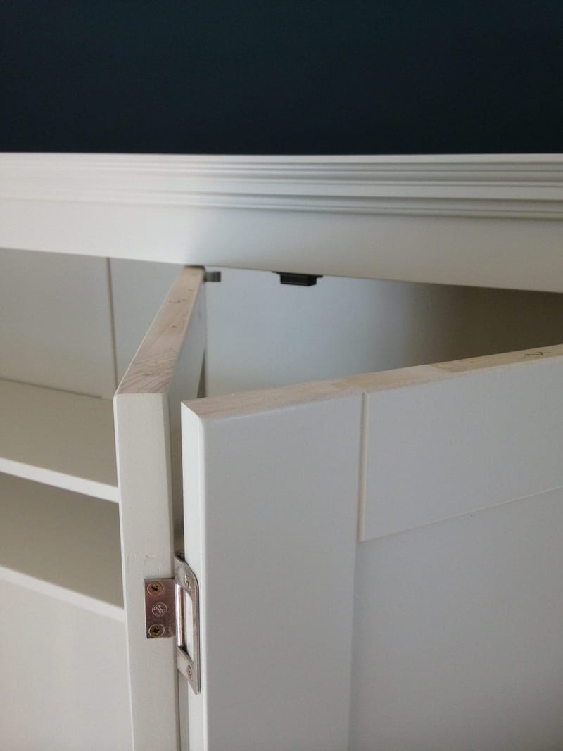 Detail of the top of a bifolding door swung open, showing the bare timber top edge, the steel butt hinge with brass screws and the moulded cornice against the dark blue wall