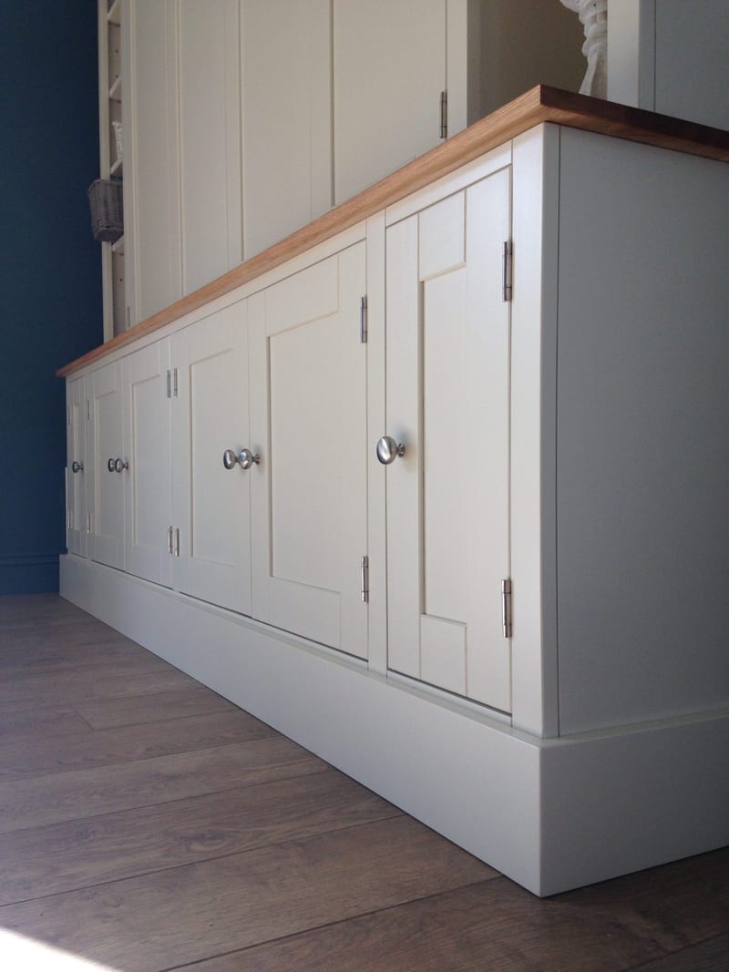 Low angled view along the base cupboards from the right end, showing the painted shaker doors, round brushed nickel knobs, steel hinges and the deep plinth on oak floorboards