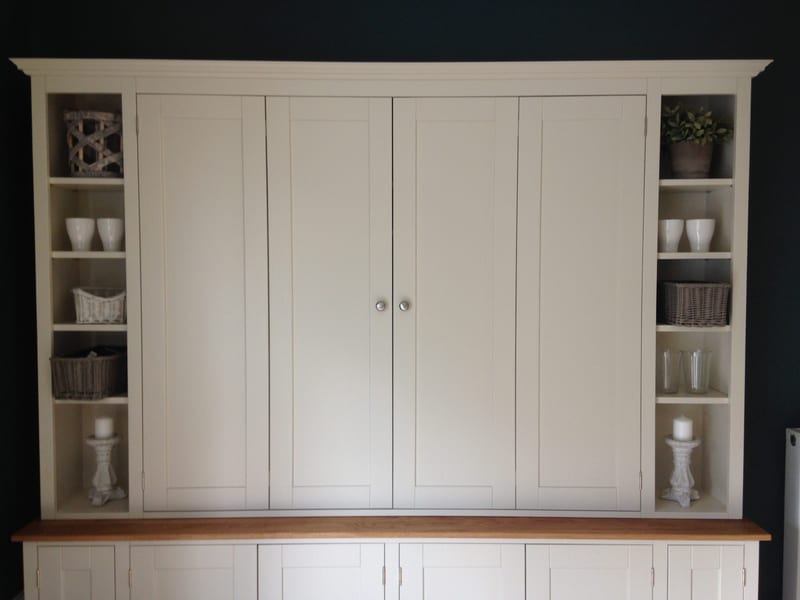 Front view of the upper section with the bifolding doors closed, flanked by open end shelves holding wicker baskets, white cups, glass tumblers and candles on stone candlesticks