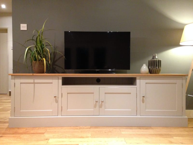 Front view of the grey painted media unit, two wide end cupboards, two centre doors and an open shelf below the television, under a light oak top against an olive grey wall