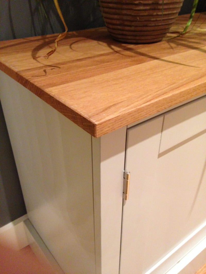 Close view of the corner of the light oak top, showing the grain and sawn edge beside the woven pot of a spider plant and the brass hinge on the grey painted end cupboard