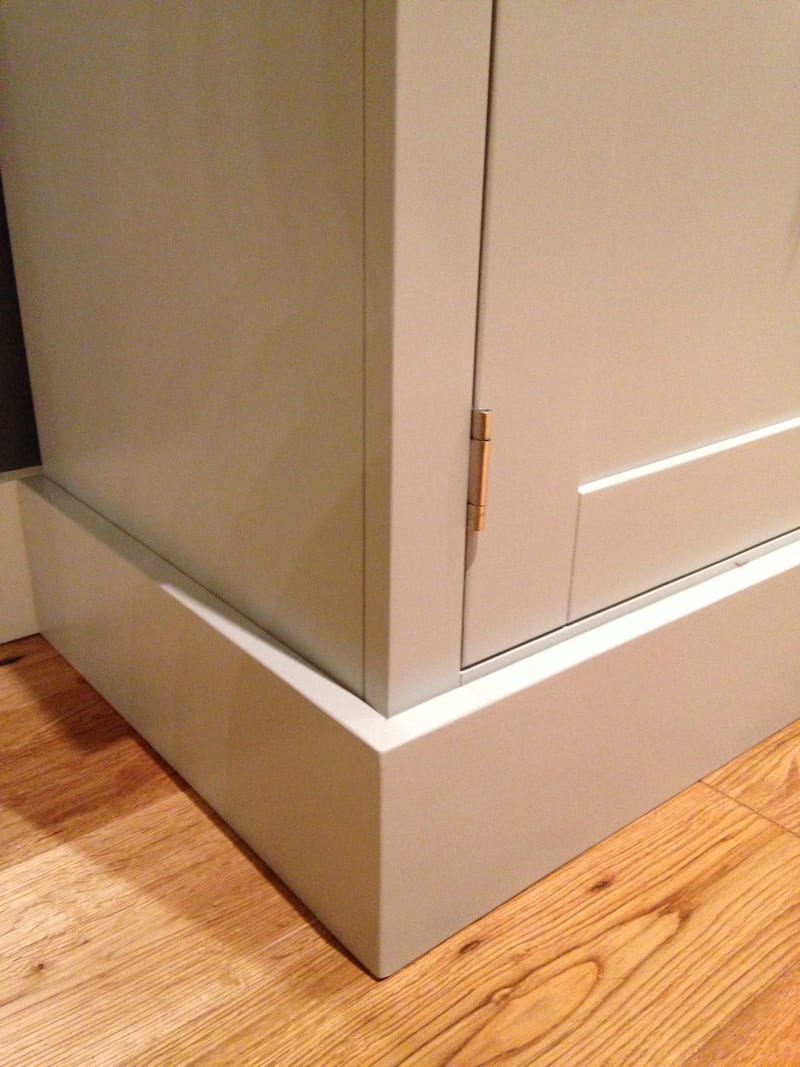 Detail of the bottom corner of the grey painted media unit, showing the plain side panel, a brass butt hinge on the shaker door and the stepped plinth resting on oak floorboards