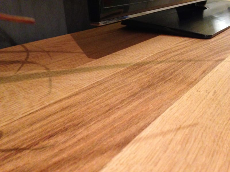 Extreme close-up across the oiled oak top, showing the fine grain and the joint between boards with the foot of the television stand at the back edge in low lamplight