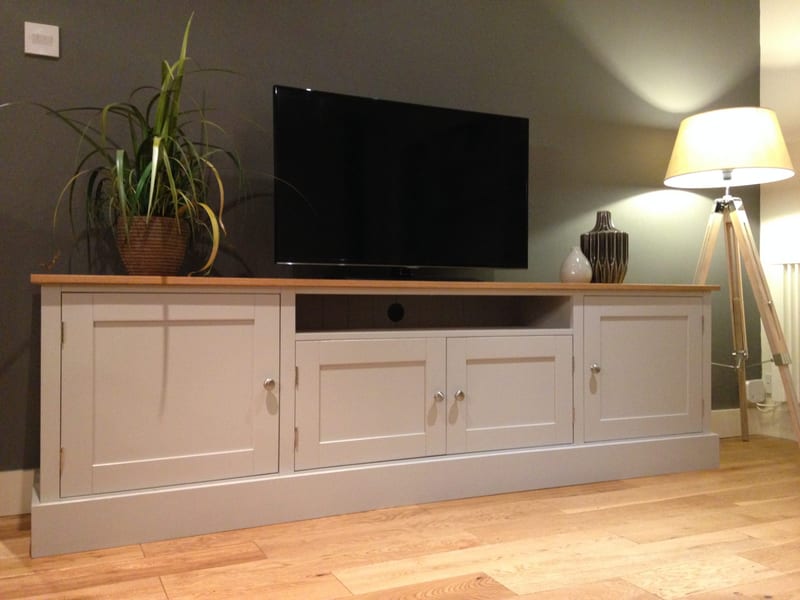 Angled full view of the grey painted media unit in a lamplit room, with a television and a potted spider plant on the oak top and two vases beside a tripod floor lamp