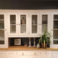 Front view of the glazed upper section, six glazed doors revealing wicker baskets, green glass bottles and a stoneware flagon above the open shelf holding candlesticks and a spider plant