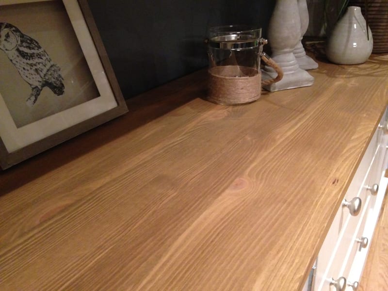 Close view along the pine top, showing the knots and grain beside a framed owl print, a rope-wrapped glass lantern, stone candlesticks and the brushed nickel drawer knobs at the end