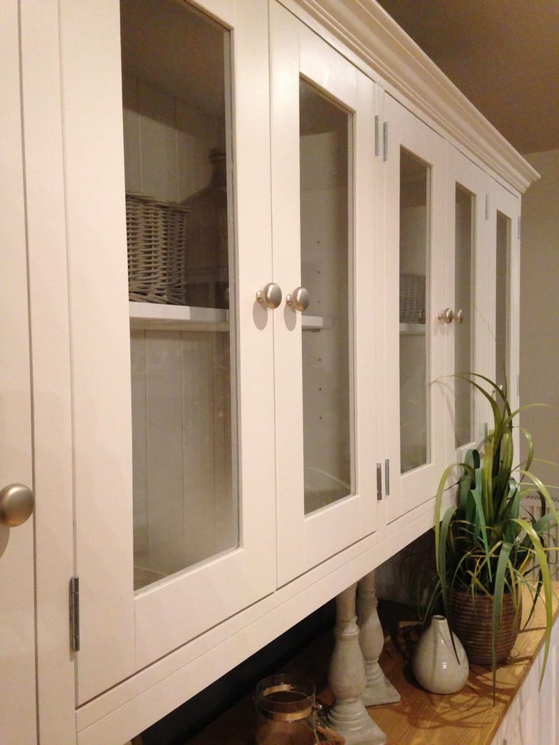 Angled close view along the glazed doors of the top, showing the shelves and wicker basket behind the glass, brushed nickel knobs and the plant and candlestick on the pine top below