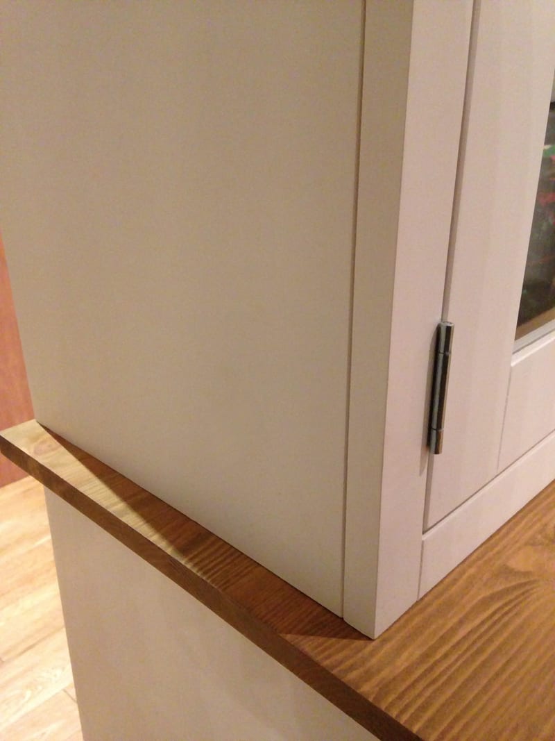 Detail of the corner where the painted top section meets the pine worktop, showing the plain side panel, the sawn edge of the top and a steel butt hinge on the glazed door